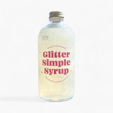 A clear glass bottle of Glitter Simple Syrup with a silver metal cap, featuring a pink and yellow label, set against a plain white background.
