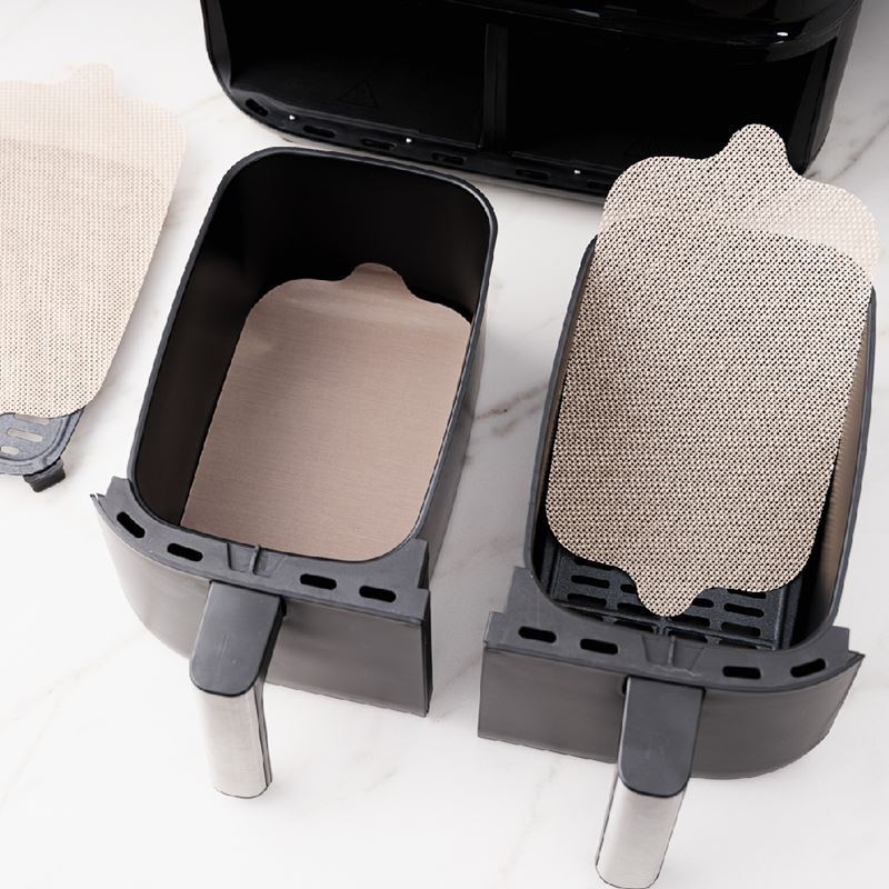 A set of four beige dual zone air fryer liners placed inside an air fryer basket, with one liner partially removed to show the textured surface.