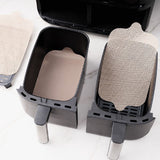 A set of four beige dual zone air fryer liners placed inside an air fryer basket, with one liner partially removed to show the textured surface.