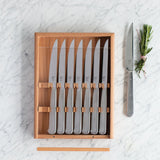 Steak knifes in a wooden storage box with one knife out on a marble counter 