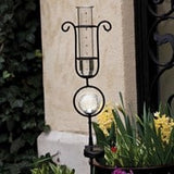 A black rain gauge with a solar light on top, placed on a stone column outdoors, with green plants around.