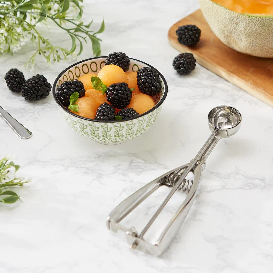 A silver-colored scoop with a spring release mechanism, resting on a marble surface with a bowl of fruit salad and a few blackberries nearby.
